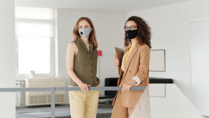 Two employees standing in a workplace, wearing masks. They are dressed work casual, and one is holding a folder.