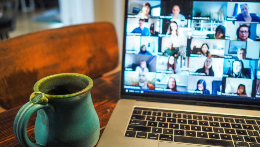 A laptop open on a desk with a coffee cup beside it. The laptop shows a meeting with a number of faces present on Zoom.