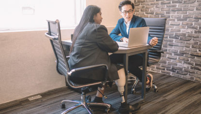Two people of colour wearing business formal clothing are seated at a desk. One with their back to the camera has a prosthetic leg. The other is working a laptop they're both looking at.