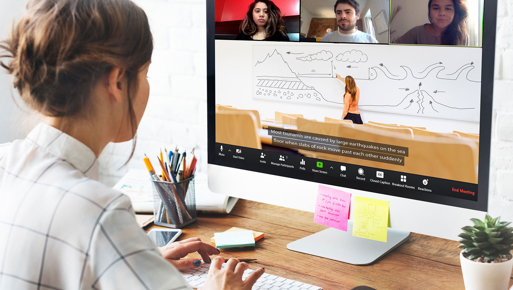 Image of a business person sitting at a desk in front of a computer monitor and typing on a keyboard, wearing a white top with black squares and their hair in a bun. They are typing captions into a video meeting with 3 people watching a presenter on Zoom.