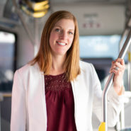 Headshot of Erinn Pinkerton. She is a white woman with light red hair. She is standing in a bus, holding a guide bar, wearing a red blouse and white blazer.