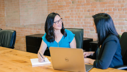 Two people of colour with shoulder length black hair are seated side by side at a wood table, behind a laptop. They look mid-conversation in a business setting.