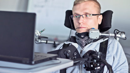 A power wheelchair user in business attire. They are using a variety of assistive technologies to work on a computer, which is positioned in front.