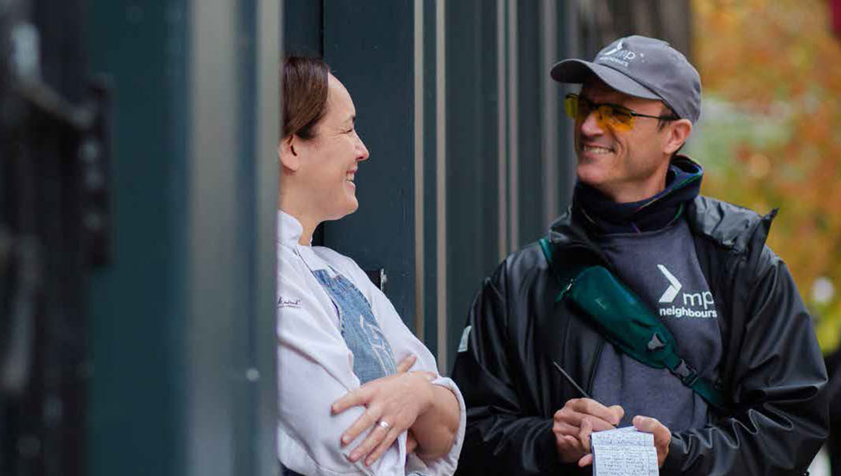 Two employees stand outside. One is wearing a blue apron, the other is wearing a Mission Possible jacket and carrying a note pad.