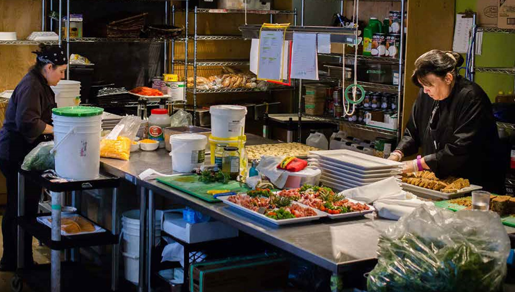 Two catering staff are preparing meals in a commercial kitchen for Potluck Cafe Society.