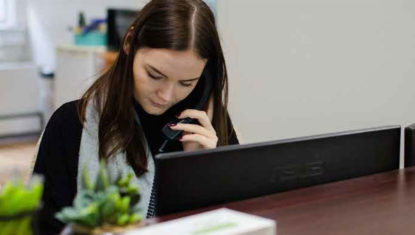 A receptionist on the phone while in front of a computer. She has dark hair and is looking down.