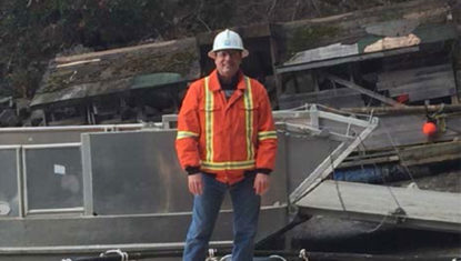 Mike is pictured wearing an orange safety jacket and white helmet in a construction zone.