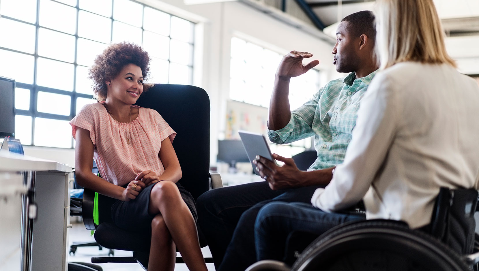 Three colleagues in an open plan workspace, conversing in a circle. Two are people of colour, and one is a white person in a manual wheelchair. They are all wearing casual business attire.