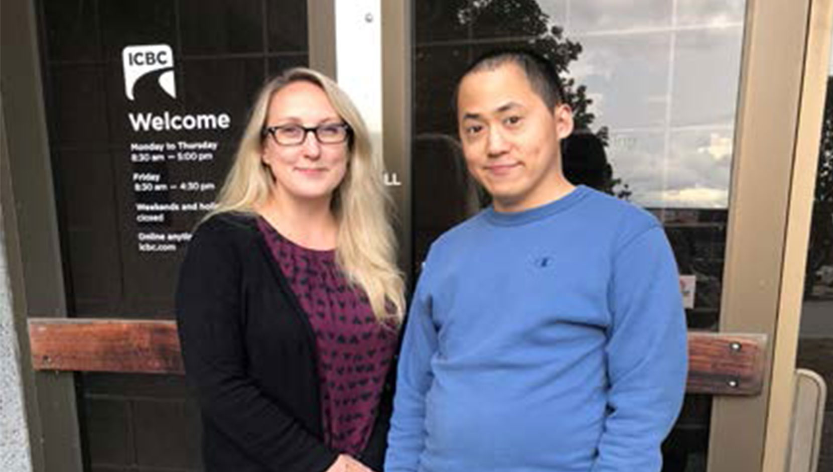 Calvin stands outside an ICBC office with his manager. He is a man of colour with short, dark hair, wearing a bright blue sweater. She is a white woman with long blonde hair and glasses, wearing a black sweater.
