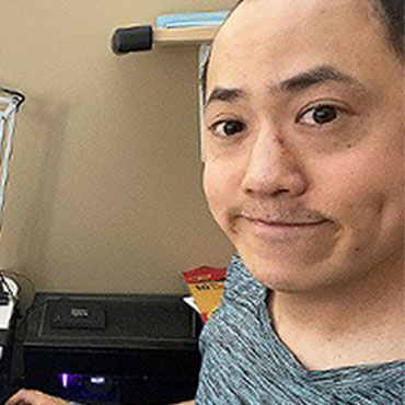 Selfie of Calvin Ng at his ICBC desk. He is an asian man with dark brown hair, slightly smiling, and wearing a grey tshirt.