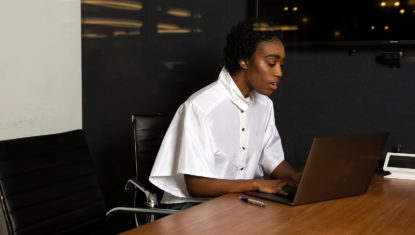 A black nonbinary person is seated at a work table with a laptop in front of them and white display board behind them. They have short black hair and a white blouse on. They're looking into a laptop and speaking during an interview.