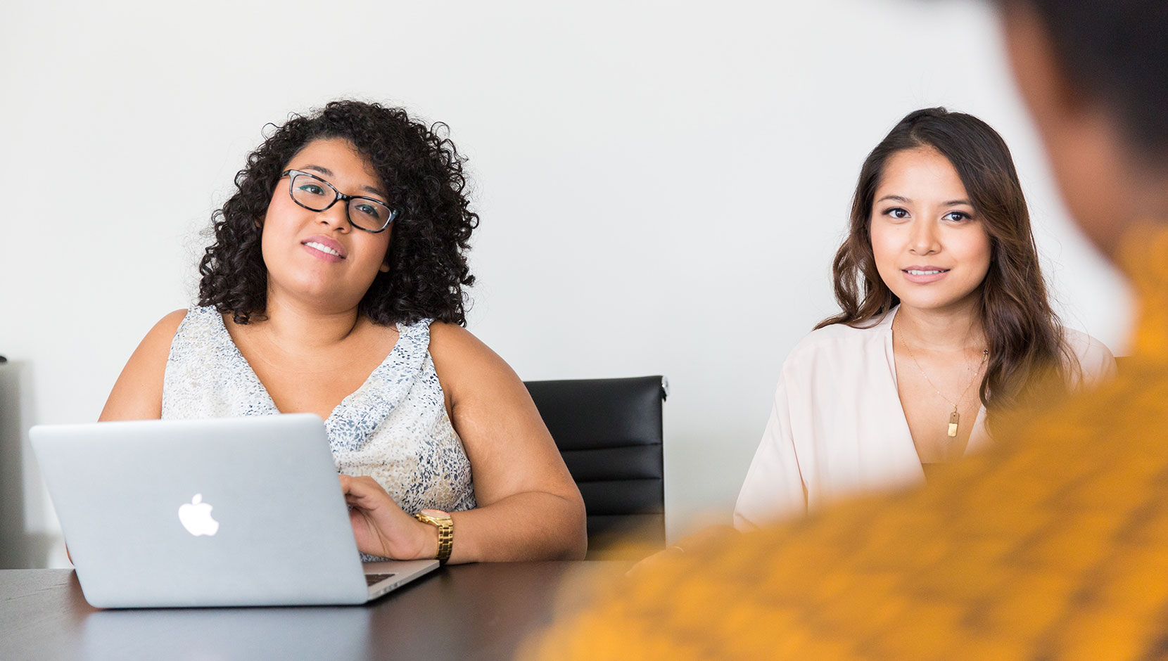 Two women of colour are facing the camera. Both are wearing business atire with shoulder length hair, and the person on the left has an open laptop. Someone's silhouette is right in the edge of the image, facing them.