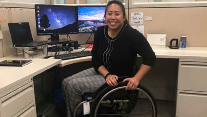 Amanda is seated in a manual wheelchair smiling in front of a desk set up with multiple screens.