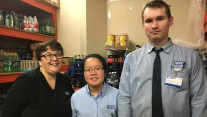 Erma, Megan and Maxwell stand in the stock room at London Drugs. Megan and Maxwell are wearing blue uniforms, and Erma is wearing a black one. They are all smiling.