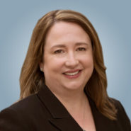 Headshot of Marilyn Tyfting. She is wearing a black blazer and has shoulder length brown hair. She is against a blue background.