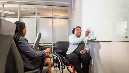 Two people of colour are working in an office meeting room. One is in a manual wheelchair, and writing on a white board. The other is facing them, watching.