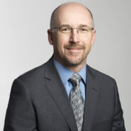 Pat Davis of BCLC. He is a white man with receeding grey hair and glasses. In this business headshot, he is wearing a dark grey suit with a light blue shirt.