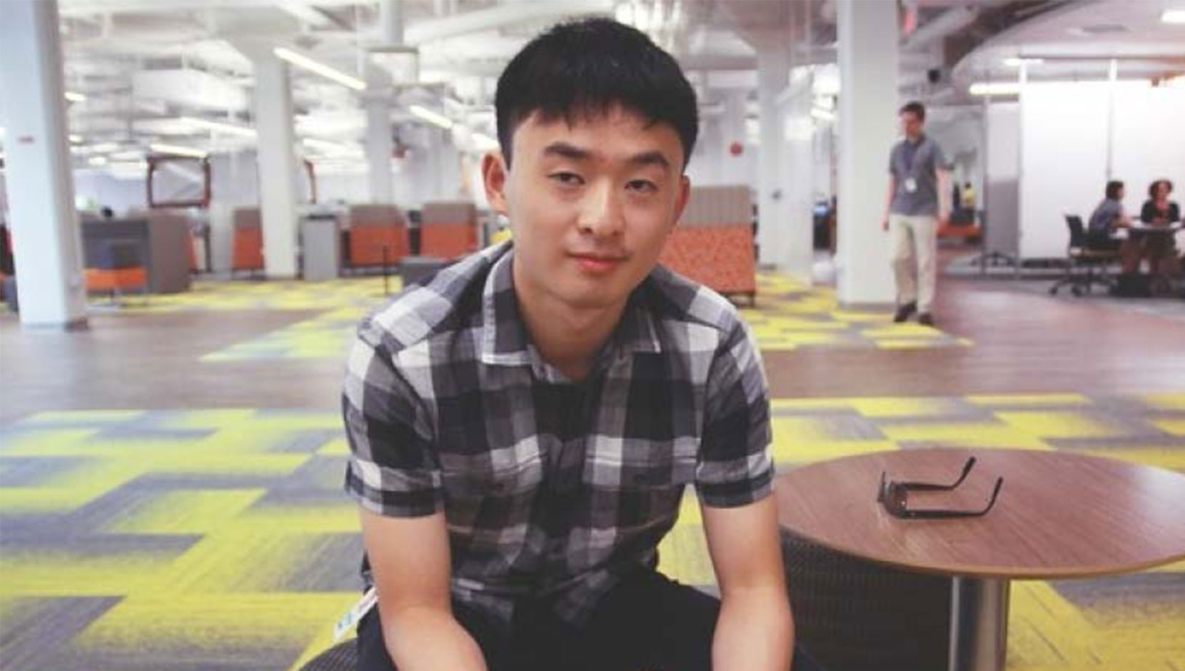 Matthew Huang of SAP. He is seated on a round chair at SAP's Vancouver office, with colourful patterned floor all around. He is an Asian man with short dark hair, and a short sleeve top.