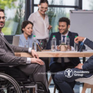 A business meeting is taking place. A variety of people are gathered around a table mid-discussion. A person in a manual wheelchair is slightly turned away from the others and smiling at the camera.