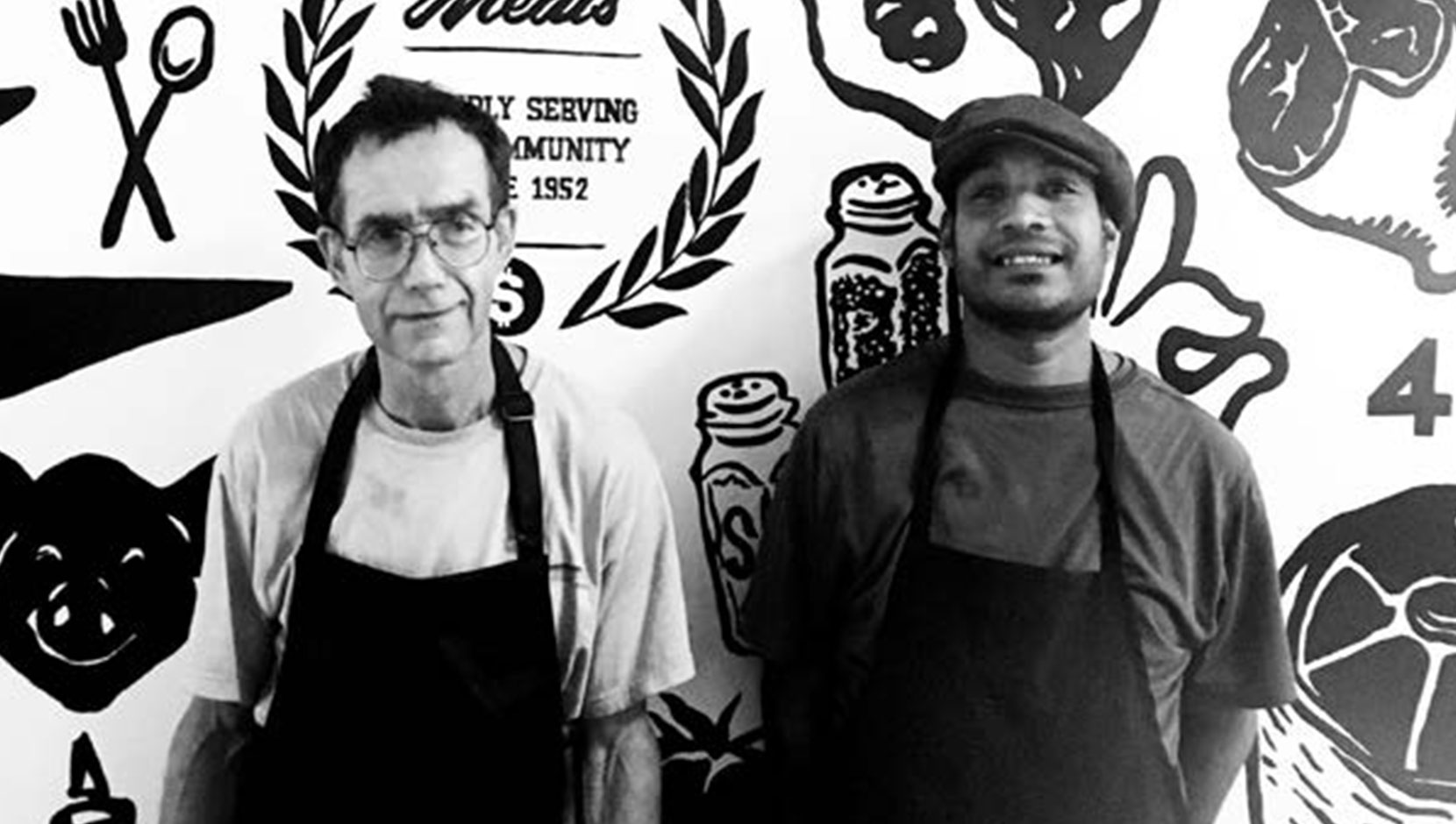 Brad and Randy leaning on a wall inside Save on Meats with an artistic food-related pattern. They're both older men and smiling. Image is in black and white.