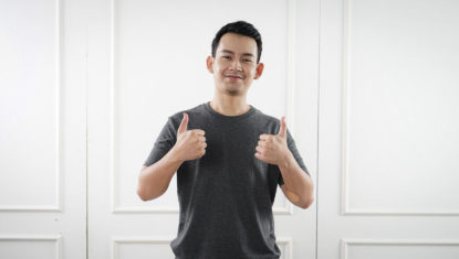 A person of colour is standing against a white background, wearing a black tshirt. They have both hands raised in a 'thumbs up' and are smiling.