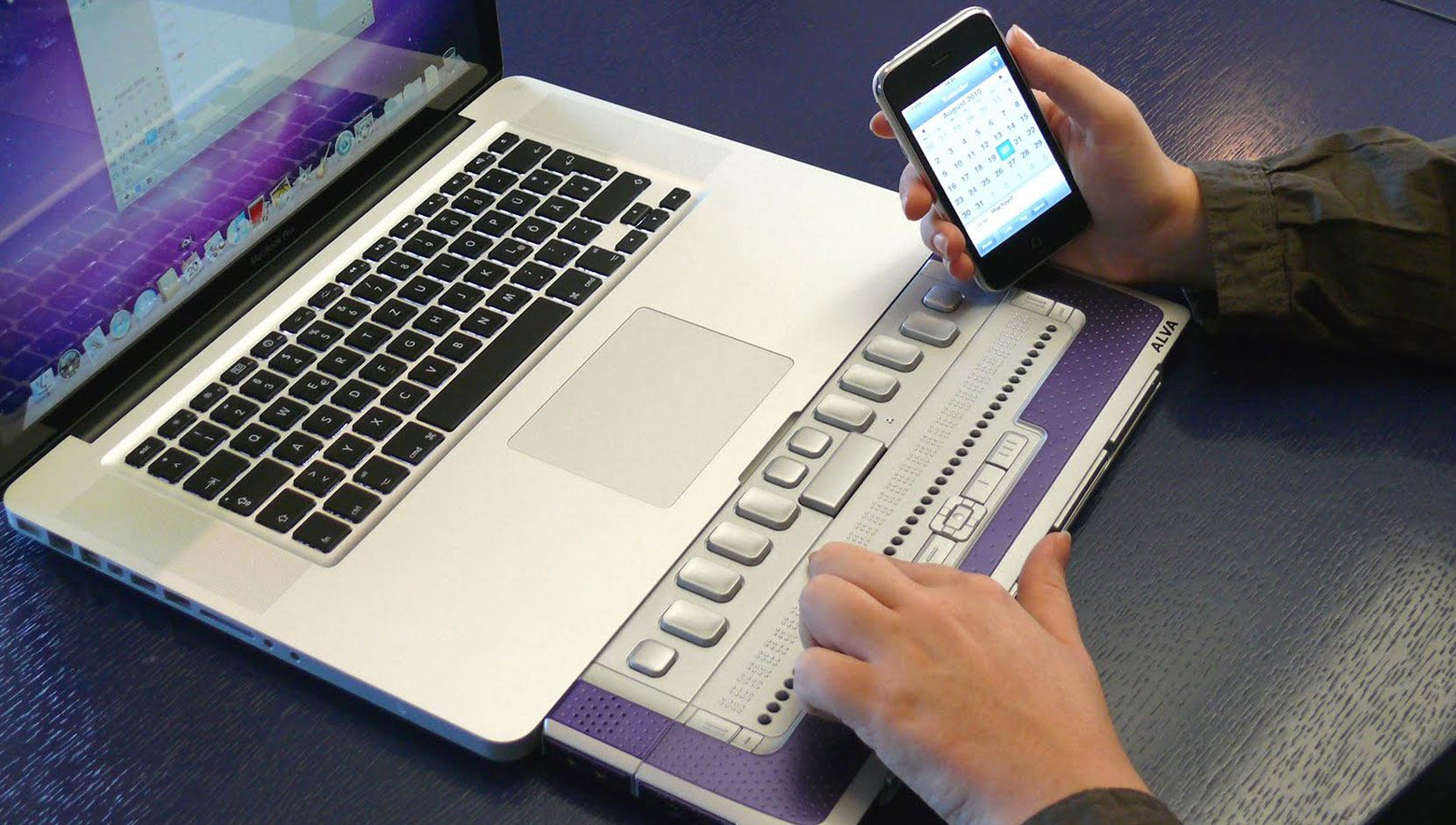 A person using a Mac laptop with a refreshable braille keyboard attachment. The person is reading the braille, with an iphone in their other hand.