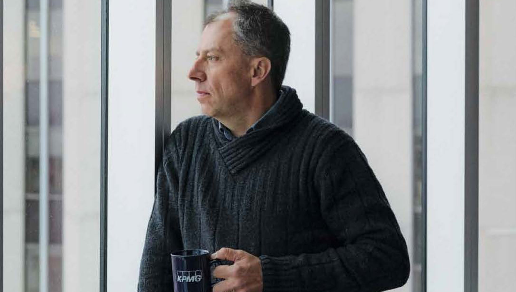 Dennis Trottier stands against floor to ceiling windows in a KPMG office. He is wearing a dark sweater, gazing outside and holding a KPMG mug.