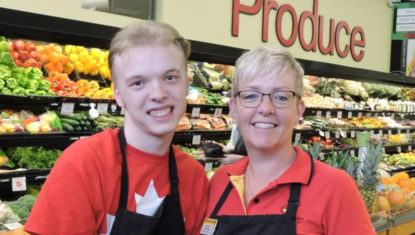 Tyler and his manager Pauline stand in the produce section of the Hope BC Buy-Low Foods, both wearing red uniforms and black aprons and smiling.