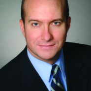 Headshot of Sam Whittaker. He is wearing a black blazer, a blue shirt and a striped blue tie.