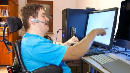 A white wheelchair user is seen in a power chair with custom seating. They are holding up their right arm and pointing at their computer screen, while talking into a headset in a home office.