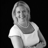Headshot of Caroline Tose of HSBC in black and white. She is visible from the waist up with her arms crossed. She is smiling, wearing a white blouse and necklace. She has short blonde hair and is against a black background.