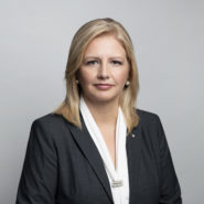 Headshot of Tamara Vrooman. She is wearing a black blazer with a white shirt. She has shoulder length blonde hair and is wearing bright lipstick, against a grey backdrop.
