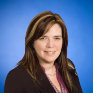Headshot of Jodi T. Kelleher. She has dark brown hair and is wearing a black blazer on a blue background.