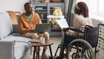 Two people of colour are facing each other in a workplace. One is in a manual wheelchair facing away from the camera, the other is facing the camera holding a laptop and seated on a couch.