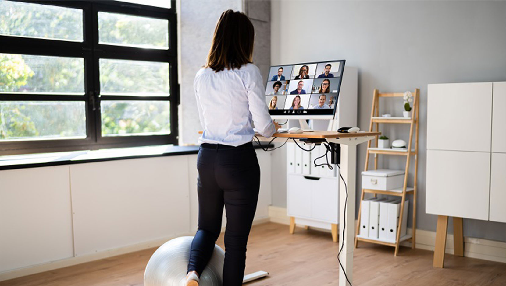 A person with dark shoulder length hair is participating in a virtual meeting, white resting their left leg on a ball at a standing desk.