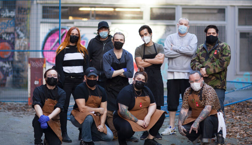 A group of 10 staff members of Save on Meats in an alleyway. They are a diverse group wearing casual clothing and aprons, with face masks.