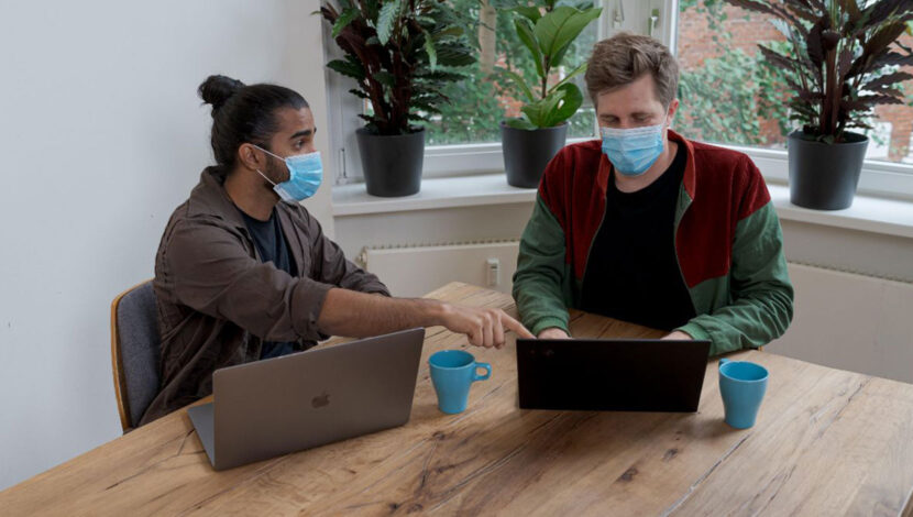 Two employees are seated at a wooden table with laptops in front of them. Both are wearing masks, and 1 is pointing at the other's screen.