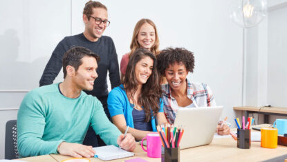 A group of 5 diverse people are gathered around a laptop and smiling.