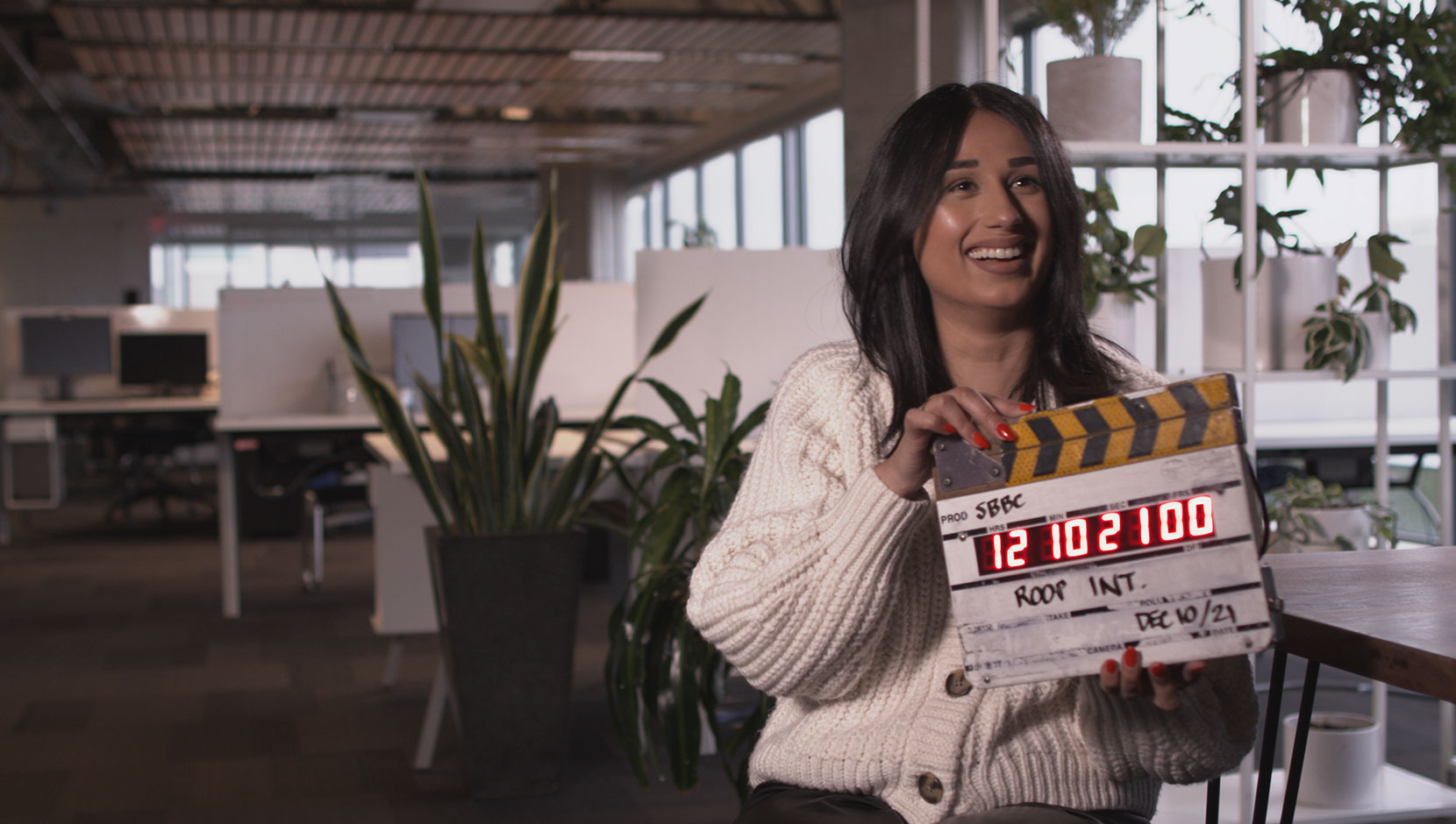Roop Johal of Small Business BC is smiling and holding a film slate. She is a woman of colour in the SBBC office, with dark hair and a cream sweater.