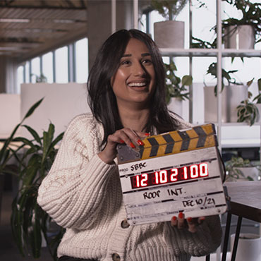 Roop Johal of Small Business BC smiles big at the camera while holding a film board during the filming of her story with True Calling Media.