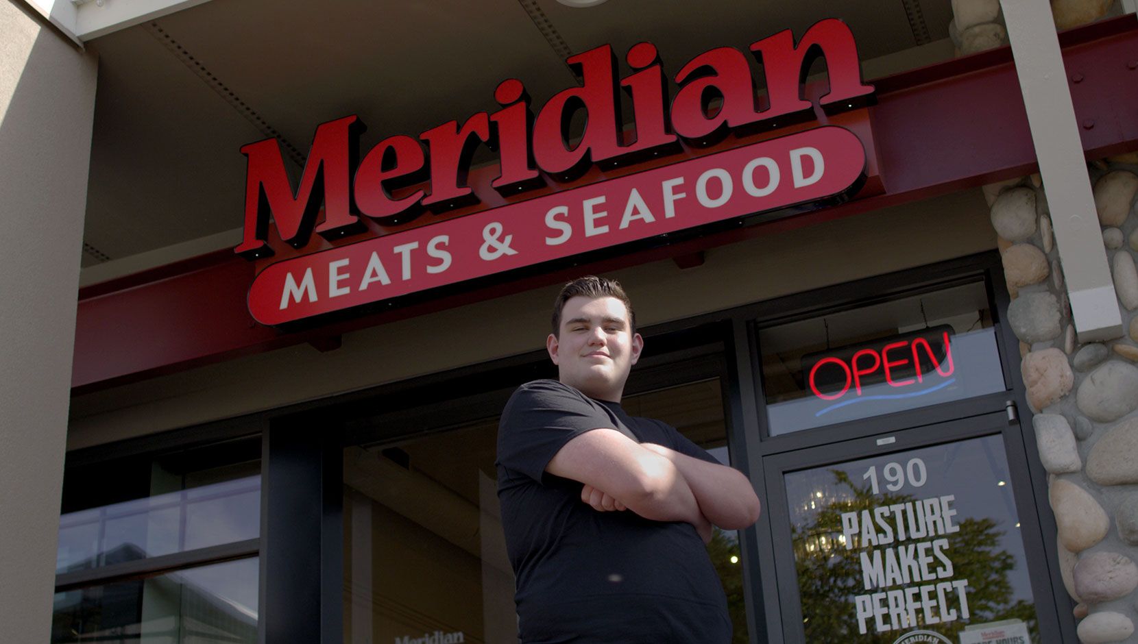 Lucas of Meridian Meats & Seafood outside of their store. He is a white high schooler with his arms crossed on his chest with a small smile.
