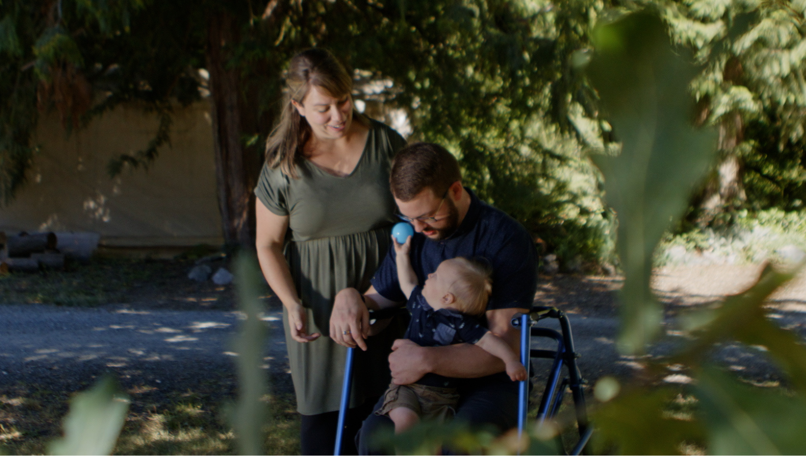 Adam Bishop with his family. The three people are gathered around his blue walker. His baby son is in his arms, reaching a ball up to his wife, standing above.