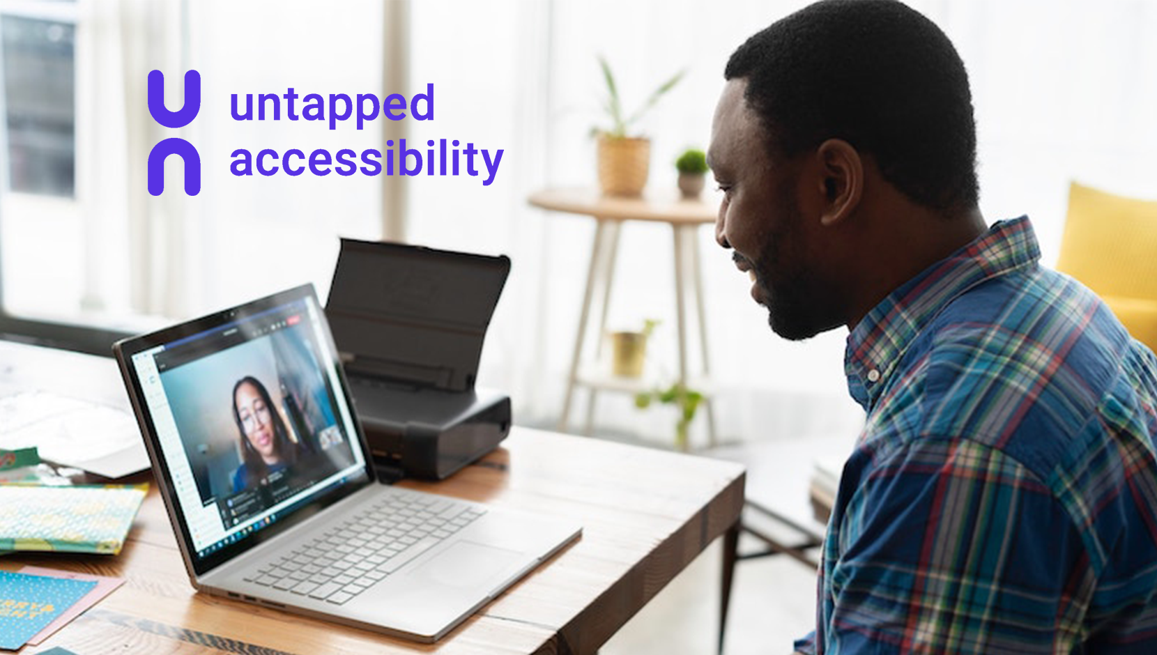 A person of colour in a virtual meeting on their laptop. They're sitting at a table in a sunny office space with the laptop open in front of them. Untapped Accessibility logo.