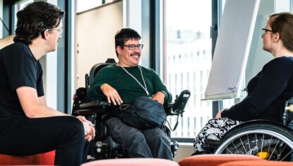 Three people with visible disabilities in conversation. The person on the left has a hearing aid, the person in the middle uses a power wheelchair, and the person on the right is in a manual wheelchair.