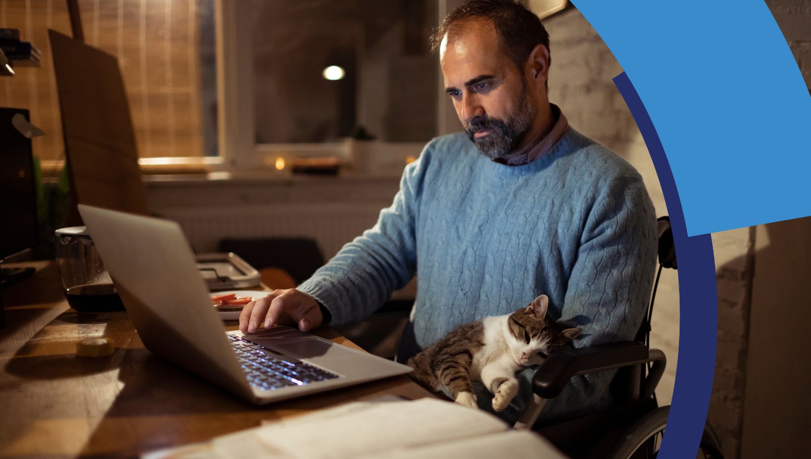 A person in a manual wheelchair is working on their laptop at a desk, with a cat resting on their lap.