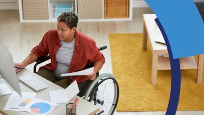 A person of colour in a manual wheelchair is sitting at a workstation reviewing pie graphs, taking notes and doing work on a computer.