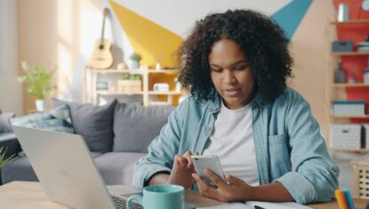 A woman sits at a desk in a bright living room, looking at her smartphone while working on a laptop. A coffee mug, notebook, and pens are on the table and colourful geometric wall art in the background.