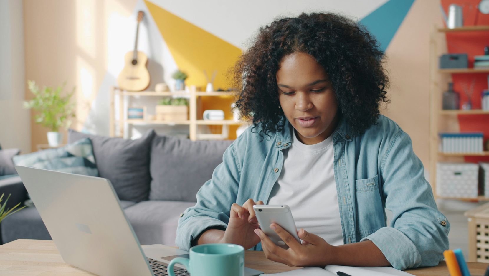 A woman sits at a desk in a bright living room, looking at her smartphone while working on a laptop. A coffee mug, notebook, and pens are on the table and colourful geometric wall art in the background.