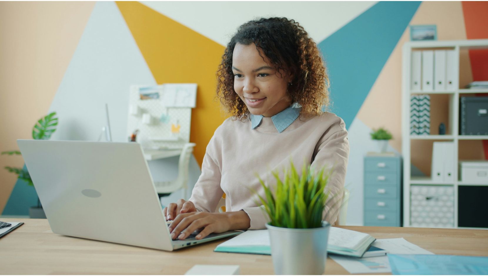 A person of colour works at a laptop in a workspace with a brightly decorated background.